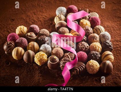 Heart shaped layout of luxury chocolate candies with pink silk ribbon over cocoa powder surface, viewed in close-up from high angle
