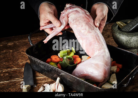 Hands of a chef preparing a wild rabbit venison roast displaying the skinned and cleaned carcass above a roasting pan with leeks, carrots and garlic o Stock Photo