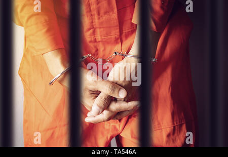 a prisoner hands in handcuffs behind the bars of a prison in orange jumpsuit clothes Stock Photo