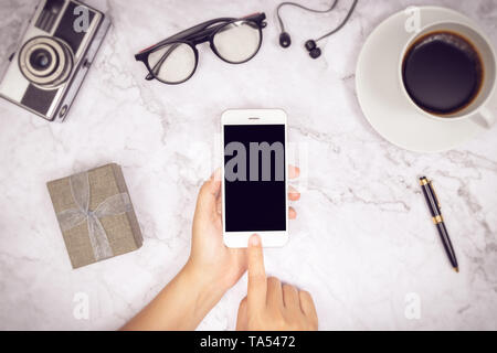 woman hand use mock up of mobile phone blank black screen with finger on touch screen with earphone,  pen, camera, glasses , cup of coffee and gift bo Stock Photo