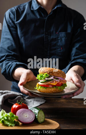 Sandwich burger with ham, cheese and vegetables on a plate in man's hands. Copy space. Fast food for breakfast or lunch. Concept for banner or adverti Stock Photo