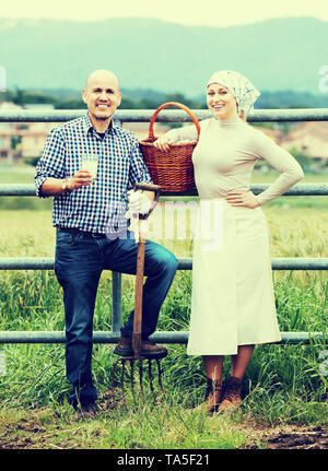 Smiling mature man and young woman standing with field fence and drinking milk. Focus on man Stock Photo