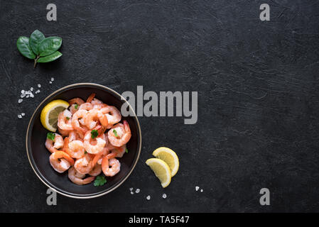 Shrimps, Prawns in bowl with lemon, sea salt and herbs, top view, copy space. Fresh seafood ingredient - shrimp tails on black. Boiled prawns. Stock Photo