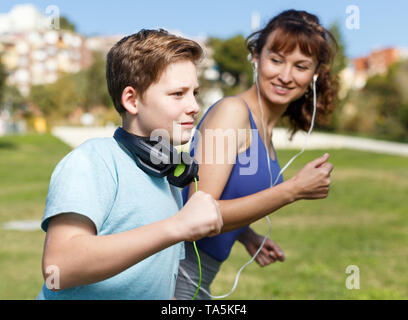 Active family of young mother and tweenage son running together on sports ground and listening music Stock Photo
