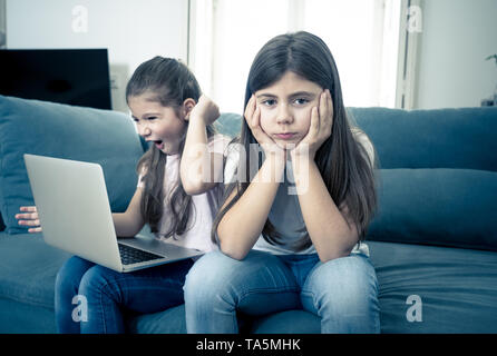 Sibling angry at younger sister spending too much time online using laptop. Digital technology addicted kid playing with computer ignoring her sad old Stock Photo