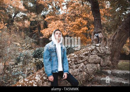 Stylish teenag boy 16-18 year old wearing white hoody with denim jacket posing outdoors. Looking at camera. 20s. Stock Photo