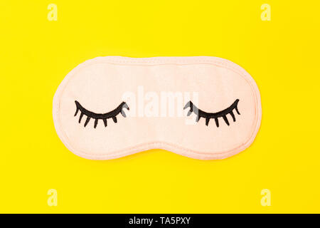Pastel pink sleep mask with closed eyes embroidered on it with eyelashes on bright yellow neon background. Fashion accessory for sleep. Stock Photo