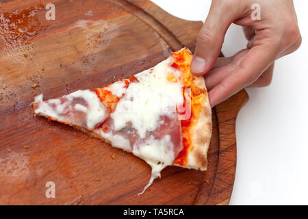 Man took last one slice of delicious Italian pizza. In frame hand taking slice of hot pizza with ham on wooden board on white table. Pizza time. Stock Photo