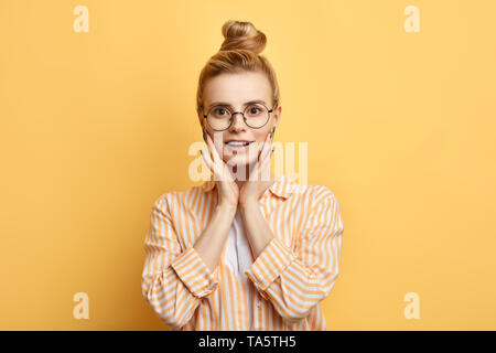 attrctive stylish female student holding palms on her cheeks and looking at the camera. close up photo. isolated yellow background Stock Photo