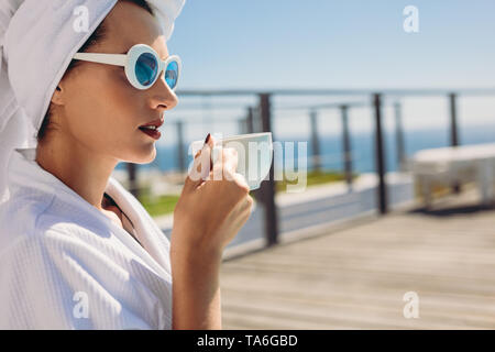 Beautiful woman sitting by pool having coffee. Female model wearing bathrobe and sunglasses relaxing at the poolside. Stock Photo