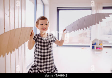 A happy small girl with cardboard paper wings playing at home. Stock Photo