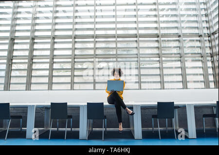 Young student or businesswoman sitting on desk in room in a library or office, using laptop. Stock Photo