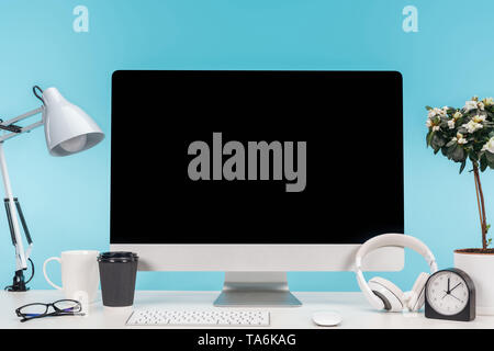 workplace with computer, lamp, cup, coffee to go, clock and flowerpot on white table on blue background Stock Photo