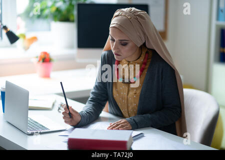 Stylish Muslim teacher sitting near laptop and correcting tests Stock Photo