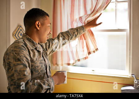 Smiling young adult soldier holding a mug while looking out the window of his home. Stock Photo