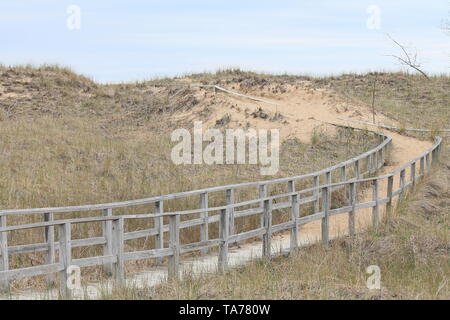 Old, weathered boardwalk cutting a path through grassy sand dunes. Sand covers much of the path Stock Photo