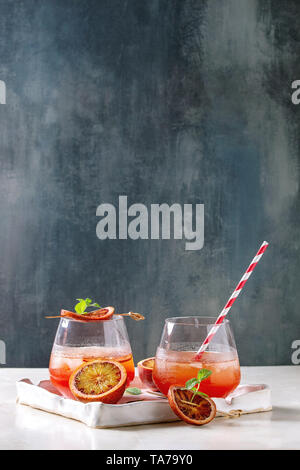 Blood orange iced cocktails in glasses, decorated by slice of oranges and fresh mint, served with retro straw in ceramic tray on white marble table wi Stock Photo