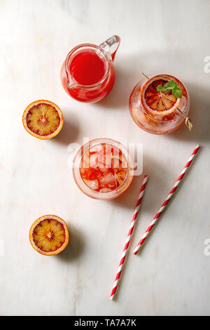 Blood orange iced cocktails in glasses, decorated by slice of oranges and fresh mint on skewers, served with retro straw over white marble background. Stock Photo