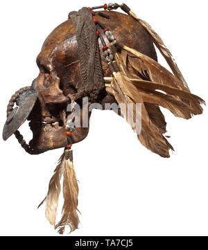 A Papua New Guinean ancestor's skull of the Asmat people Dark patinated male skull, the jaw joint wrapped with textile braid, the eye sockets adorned with plant seeds, the nasal cavity connected to the lower jaw by a band of braided plant fibers and decorated with a snail shell. Elaborate headdress made of textile, plant seeds and feathers (some feather bundles loosely enclosed). Height 21 cm. historic, historical, Indonesian archipelago, Indonesia, Far East, Asia, Asian, ethnology, ethnicity, ethnic, tribal, object, objects, stills, clipping, cl, Additional-Rights-Clearance-Info-Not-Available Stock Photo