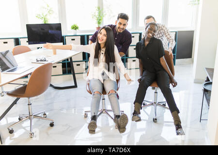 Carefree excited diverse workers having fun riding oh chairs celebrating friday together, happy employees enjoy funny competition laughing together fe Stock Photo