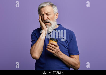 Tired exhausted old man with sleepy expression, covers face with palm, holds disposable paper cup of coffee.close up portrait. Stock Photo