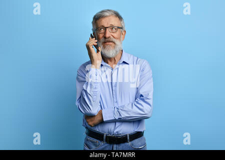 handsome senior man looking up, talking on mobile phone isolated against sky blue background. old businessman having chat, business conversation witj  Stock Photo