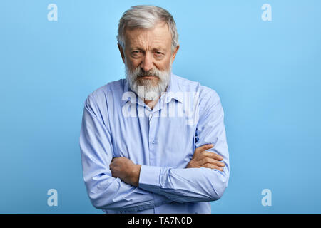 Handsome senior man in blue shirt with skeptic, nervous expression standing with crossed arms. close up portrait. isolated light blue background Stock Photo