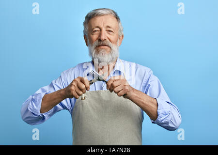 old cheerful chef or waiter in gray apron, blue shirt holding fork, spoon isolated on blue background. houseworker, domestic worker.crossed cutlery Stock Photo