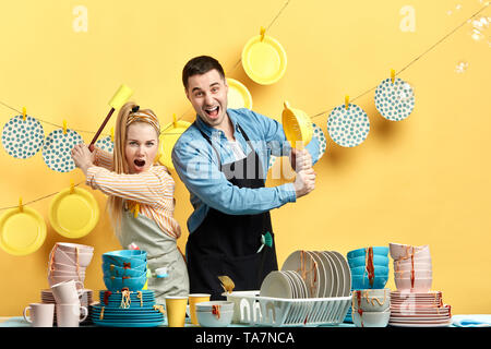 Funny positive couple in aprons having fun during doing housework and cleaning. crazy man and woman fighting with dirty cutlery.we're going to wash yo Stock Photo