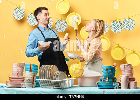 funny young blonde woman with closed eyes blowing foam to her boyfriend in the kitchen, side view photo. happy pastime concept Stock Photo
