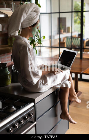 Woman in bathrobe sitting on the kitchen counter and using laptop Stock Photo
