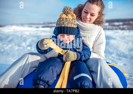 A young mother, woman with child, boy, son of 3 years old, in the winter outside in warm clothes, sitting on a tubing, rolling down a hill, playing Stock Photo
