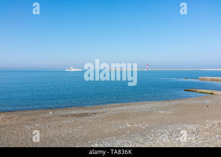 A large white yacht enters the port. Deserted pebble beach. Clear blue sky over the seaport. Stock Photo