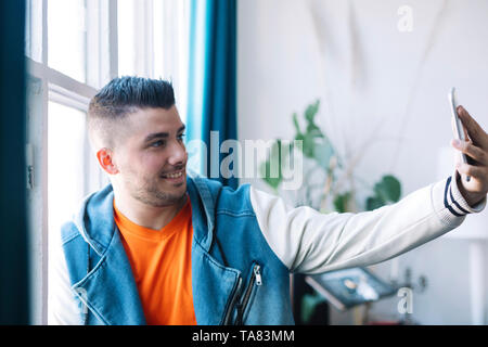 A Smiling Handsome Young Man Taking Self Picture Using Mobile Phone Inside the House. Stock Photo