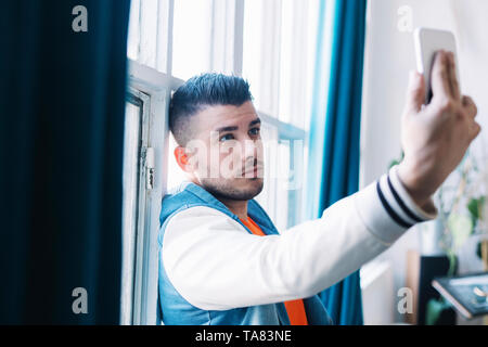 A Smiling Handsome Young Man Taking Self Picture Using Mobile Phone Inside the House. Stock Photo