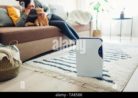 Dehumidifier with touch panel, humidity indicator, uv lamp, air ionizer, water container works at home while woman playing with cat. Air dryer Stock Photo