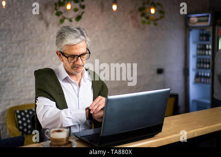 Senior man checking time while using laptop and having coffee in the bar Stock Photo