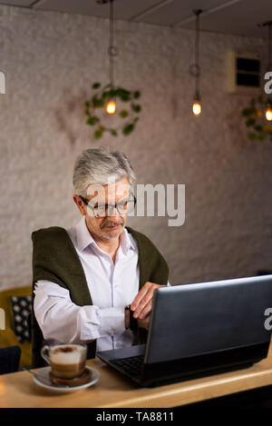 Senior man checking time while using laptop and having coffee in the bar Stock Photo