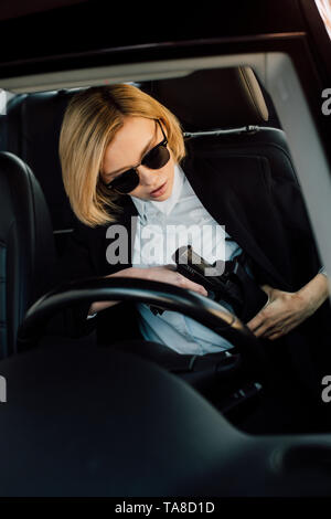 beautiful young blonde woman in sunglasses holding gun in car Stock Photo