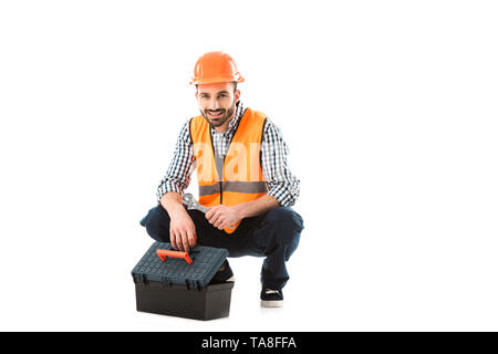 cheerful construction worker sitting near toolbox and holding adjustable wrench isolated on white Stock Photo