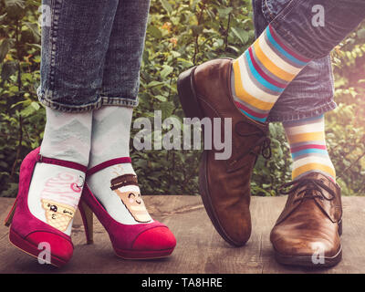 Men's and women's legs in fashionable shoes, bright, multi-colored socks on a wooden terrace on the background of green trees and sunlight. Close-up.  Stock Photo