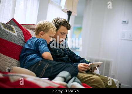Two brothers looking at an iPad together while sitting on a couch inside a living room. Stock Photo