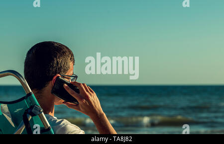 Young man, speaks by mobile phone sitting on the beach, manages his work from anywhere in the world Stock Photo