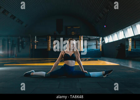 Stretching gymnast girl doing vertical split, twine. Side view of attractive young woman doing core exercise at crossfit gym Stock Photo