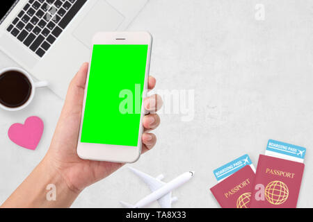 woman hand holding a modern mobile smart phone with blank green screen. two passport , coffee , computer laptop  at background Stock Photo