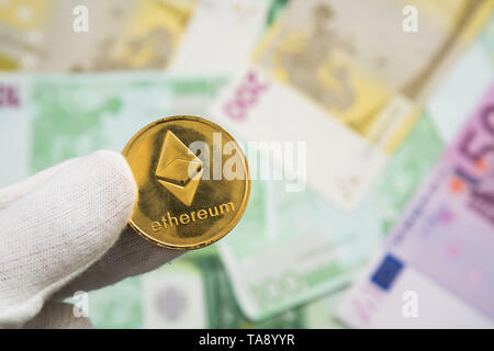 Man in white cloves holding Ethereum coin between fingers with Euro bank notes in the background. Digital currency, block chain market Stock Photo