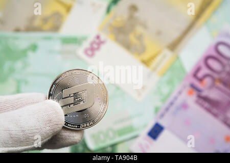 Man in white cloves holding Dash coin between fingers with Euro bank notes in the background. Digital currency, block chain market Stock Photo