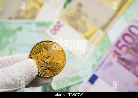 Man in white cloves holding Monerd coin between fingers with Euro bank notes in the background. Digital currency, block chain market Stock Photo
