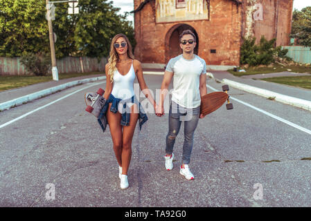 Young couple man woman walking summer city, skate longboard. Youth fashion style, modern leisure trend, tanned sports figure. Emotions of happiness Stock Photo