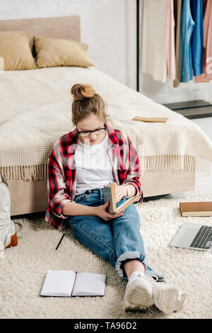 Concentrated teenager in glasses and checkered shirt reading book near bed Stock Photo